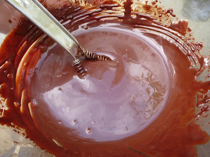 Close up detail of melted, glossy chocolate in a glass mixing bowl with a stainless steel whisk.