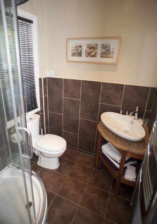 Interior of a compact modern bathroom with corner units for the shower and hand basin, brown tiles and a white toilet below a window with blind