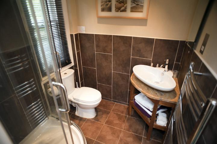 Ensuite bathroom interior with brown tiles, a corner hand basin and shower and toilet below a window with Venetian blinds