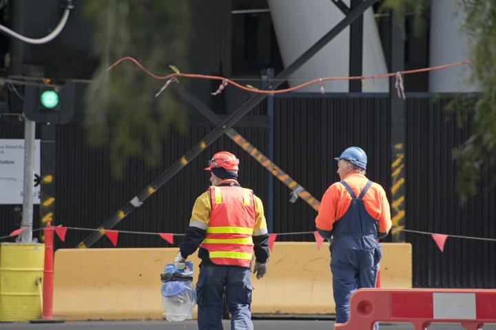 Two construction workers on a road crew standing on a building site with their backs to the camera watching for traffic with a green light in the background