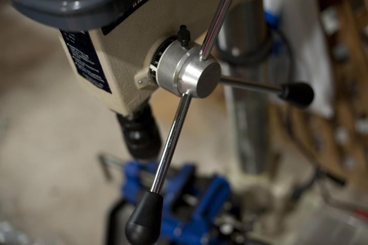 Close up of a metal drill press with winding handle in a cluttered workshop.