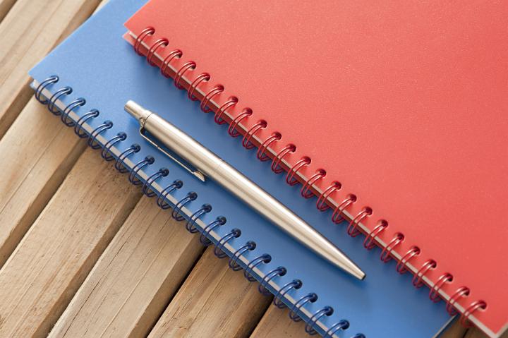 Stationery Concept - Close up Silver Ballpoint Pen on Blue and Red Spiral Notebook on Top of Wooden Table