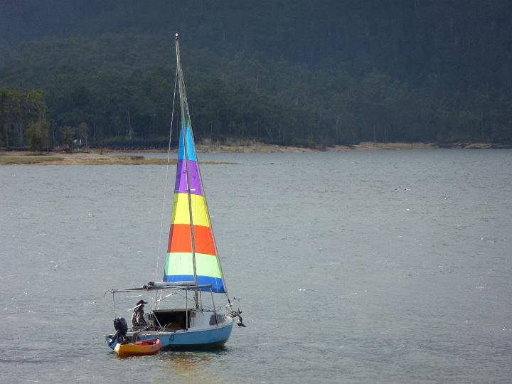 Small sailboat with a colorful sail sailing offshore with a lone sailor towing a dinghy for access to the shore from a mooring