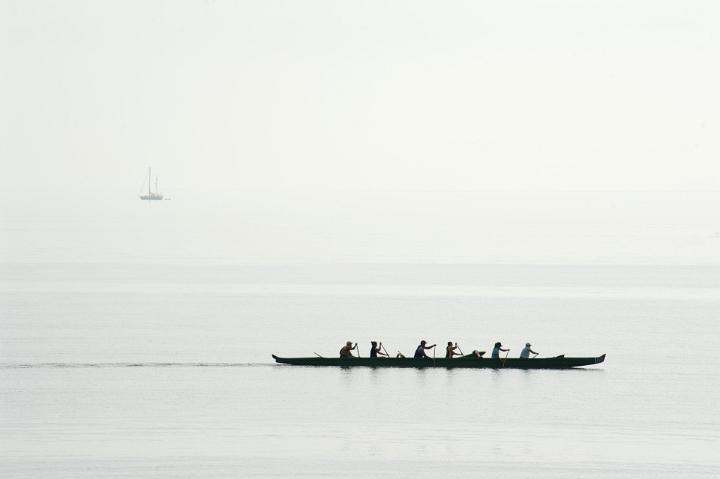 Six men team training for a race in their canoe paddling in unison on a calm ocean, silhouette against a misty sea