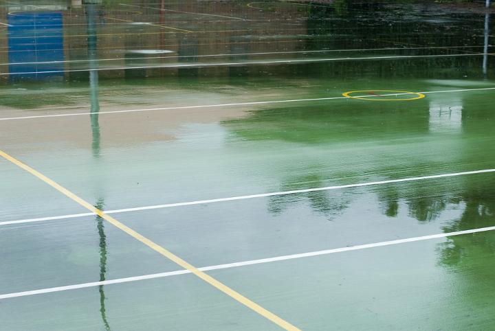 Wet outdoor all-weather sports court with yellow and white lines on a green surface reflecting surrounding trees on the surface of the water