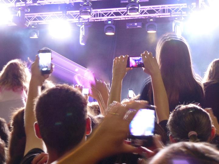 Crowd of fans at a live concert performance on a night out photographing the band on their mobile phones and cheering , view from the back