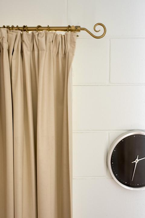 An abstract home decor with beige curtain, rod and black clock hanging on a white brick wall.