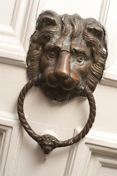 Brass door knocker of a lions head with a ring in its mouth for banging in order to gain access to a white painted front door