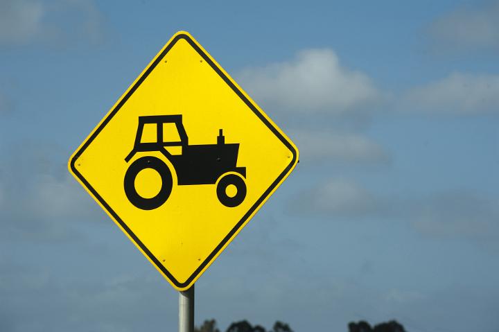 Yellow farm equipment and machinery traffic warning sign showing the silhouette of a tractor against a blue sky with copyspace