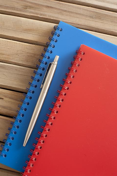 Close up Silver Ballpoint Pen and Spiral Blue and Red Notebook on Top of Wooden Table