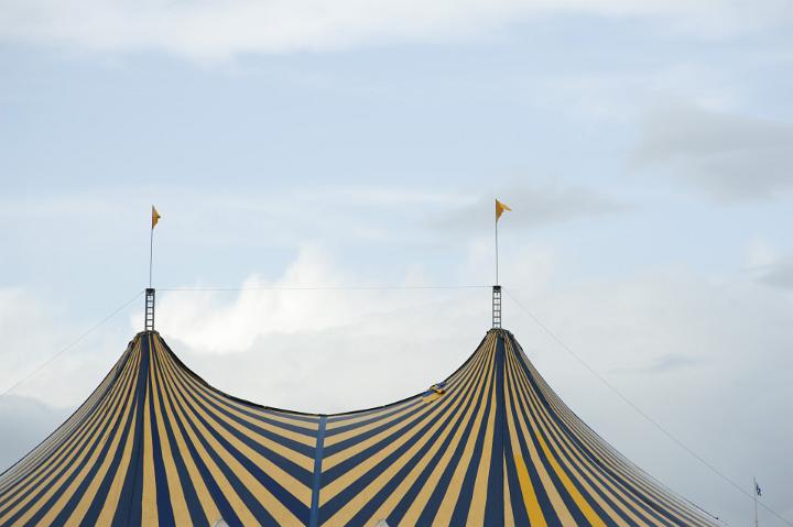 Striped Big Top circus tent with colorful alternating yellow and blue stripes and flags on the poles against a cloudy sky