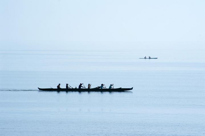 Team of rowers competing in a race in a six man canoe silhouetted against a calm ocean as they paddle furiously to gain speed