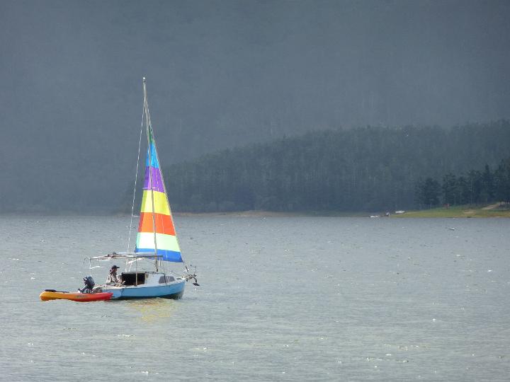 Small sailboat with a single colourful sail towing a dinghy offshore on a misty cold day with the coastline visible in the distance