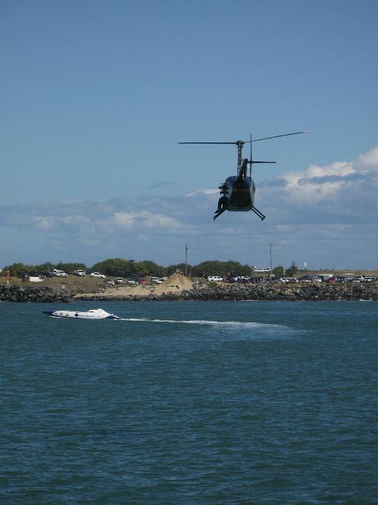 Helicopter flying above a speedboat cruising offshore with houses visible on a headland in the background