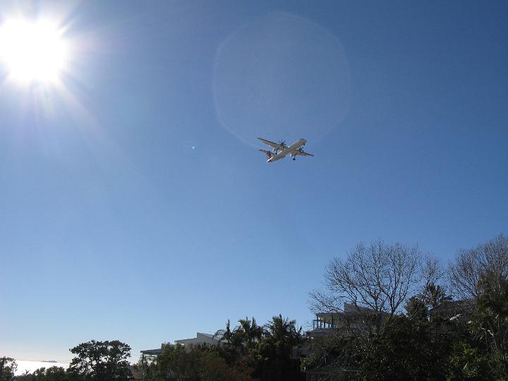 Low Angle View of Small Aircraft Flying over Hillside Homes in Large Blue Sky with Bright Sun