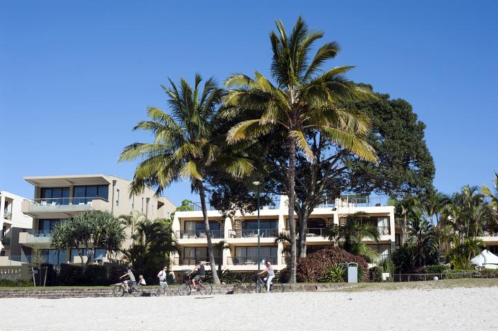 Group of People Riding Bicycles Past Low-Rise Beachfront Resort with Palm Trees
