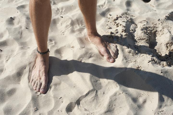 Close Up of Male Bare Feet with Ankle Bracelet Standing on Sandy Beach with Shadow from Bright Sun